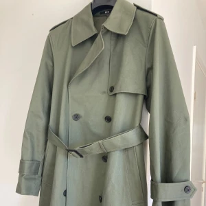 Olivgrön trenchcoat från Uniqlo - Snygg olivgrön trenchcoat från Uniqlo med klassisk dubbelknäppning, axelklaffar och bälte i midjan. Yttertyg i bomull. Perfekt för vår och höst.
