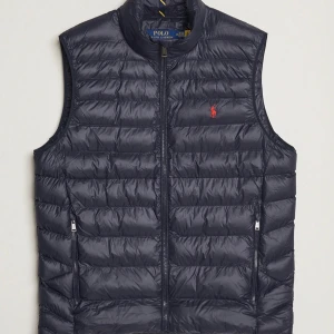 Mörkblå dunväst från Polo Ralph Lauren - Snygg mörkblå dunväst från Polo Ralph Lauren med dragkedja framtill och två fickor med dragkedja. Västen har ett quiltat mönster och en liten röd broderad logga på bröstet. Perfekt för lager-på-lager-stil. 