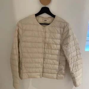 Beige lättviktsdunjacka från Uniqlo - Säljer en beige, quiltad lättviktsdunjacka från Uniqlo i storlek M. Den har en fläck som syns i vissa ljus men inget man tänker på:) Skulle tro att fläcken försvinner i Kemtvätt!! Jackan har rund halsringning, tryckknappar framtill och är långärmad. Perfekt för lager-på-lager och enkel att bära med sig tack vare sin lätta design.