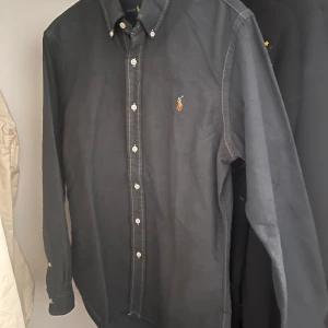Mörkblå skjorta från Ralph Lauren - Klassisk mörkblå skjorta från Ralph Lauren med knappar hela vägen och broderad logga på bröstet. Skjortan har lång ärm, button-down krage. Perfekt för en stilren look inför sommarn! Hör av för fler frågor!