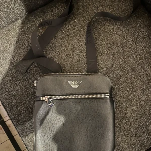 Svart axelväska från Armani - Snygg svart axelväska från Armani med justerbart band och silverfärgad dragkedja framtill. Väskan har en stilren design med logotypen i metall på framsidan och är tillverkad i ett syntetmaterial med struktur.