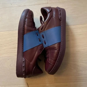 Vinröda sneakers Valentino Garavani med blå detalj - Säljer ett par vinröda Valentino Garavani sneakers med bred blå rem över mitten. Skorna har snörning och platt sula, tillverkade i skinn med klassisk Valentino-logga på sulan och insidan. Perfekta för dig som gillar stilrena och exklusiva sneakers. Finns ingen sko låda. Hör av dig vid frågor 