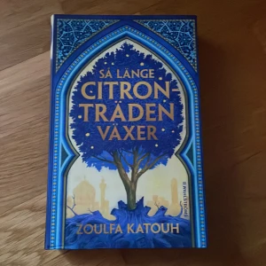 Så länge citronträden växer - Zoulfa Katouh - En roman med blått hårt omslag och guldiga detaljer. Boken är skriven av Zoulfa Katouh och utgiven av B. Wahlströms. Omslaget pryds av ett träd och silhuetter av byggnader, och berättelsen handlar om kärlek och frihetskamp i Syrien.