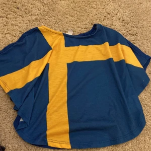 Blå och gul Sverige  t-shirt från Lindex Kids - Säljer en blå t-shirt från Lindex Kids med ett stort gult korsmönster framtill, inspirerat av svenska flaggan. T-shirten har korta ärmar och är tillverkad i mjuk bomull, perfekt för barn i åldern 6-7 år.