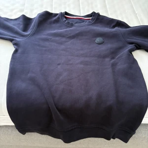 Mörkblå sweatshirt från Moncler - Säljer en stilren mörkblå sweatshirt från Moncler med rund halsringning och diskret logotyp på bröstet. Tröjan har långa ärmar och är tillverkad i ett mjukt bomullsmaterial. Perfekt för en avslappnad och snygg look.