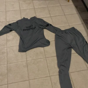 Grå långärmad PSG fotbollströja med byxa - Säljer en grå långärmad PSG-träningströja från Nike med svarta detaljer och röd Nike-logga. Tröjan har texten 'Accor Live Limitless' och PSG-emblem på bröstet. Matchande grå träningsbyxa med PSG-märke ingår.