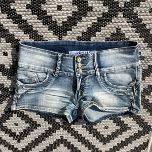 Blå jeansshorts med knappar - Snygga ljusblå jeansshorts med slitningar och fyra guldfärgade knappar framtill. Shortsen har låg midja, fram- och bakfickor med nitar och hjärtformade bakfickor. Perfekta för varma dagar och en avslappnad stil.