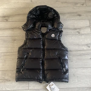 Svart dunväst från Moncler - OBS: storlek M fast passar som S Säljer en svart dunväst från Moncler med huva och två dragkedjefickor framtill. Västen har en quiltad design och stängs med dragkedja. Klassisk Moncler-logga på bröstet och märkestaggar kvar.