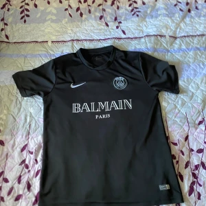 Svart PSG fotbollströja Nike x Balmain - Säljer en svart PSG fotbollströja i samarbete med Balmain och Nike. Tröjan har korta ärmar, rund hals och tydliga vita tryck med Paris-logga och Balmain Paris på bröstet. Tillverkad i ett lätt och ventilerande material, perfekt för fotboll eller streetwear.