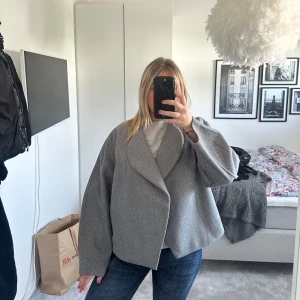 Grå oversized kappa med bred krage - Säljer en grå oversized kappa med bred, asymmetrisk krage och långa ärmar. Jackan har en stilren och minimalistisk design utan synliga knappar eller dragkedja. Perfekt för dig som gillar en modern och avslappnad look. Jackan är helt ny , med andra ord aldrig använd. Perfekt till en sommarkväll eller bara till en snygg outfit!