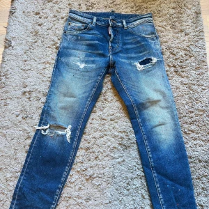 Blå slitna jeans från Dsquared2 - Säljer ett par blåa jeans från Dsquared2 med slitningar och hål på låret och knät. Jeansen har klassisk femficksdesign, raka ben och detaljer som små färgstänk. Midjemått ca 35 cm. Perfekta för en cool och avslappnad stil.