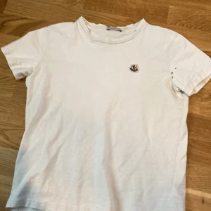 Vit t-shirt från Moncler - Säljer min vita moncler t shirt då den inte längre passar mig. Den är i ganska dåligt skick då den har mycket små hål i armhålan och på ryggen men den ör perfekt att ha som undertröja under en jacka eller hoodie som jag har gjort. Vet inte vilken storlek den är men han jag köpte den av sa xs och jag brukar ha S/M men den ör lite för liten för mig nu så tror att xs stämmer bra. Bara att höra av sig vid frågor eller bilder.