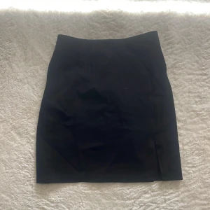 Svart pennkjol med slits - En klassisk svart pennkjol med diskret slits framtill. Kjolen har en rak och tight passform som sitter snyggt på kroppen. Perfekt att matcha med en enkel topp eller skjorta.