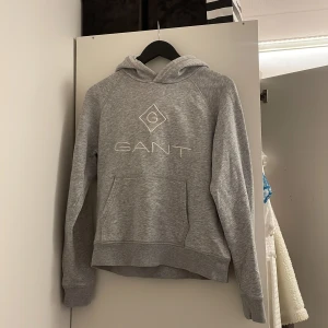 Grå hoodie från GANT - Säljer en klassisk grå hoodie från GANT med broderad logga framtill. Tröjan har huva, lång ärm och en stor magficka. Perfekt för en avslappnad och stilren look.
