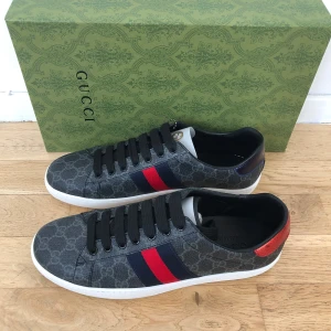 Gucci ace skor - Snygga Gucci ace skor i grått/svart med GG-monogram över hela skon. Skorna har svarta snören, vit sula och klassiska ränder i rött och mörkblått på sidorna. Baktill har ena skon röd metallic-detalj och den andra mörkblå. Perfekt för dig som vill sticka ut. Skorna är helt nya, inte använda en ända gång. Säljer dem helt enkelt för dom bara inte passar mig 