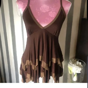 Brun topp från Zara - Supersöt brun linne från Zara med volangdetaljer och v-ringad hals. Linne har smala axelband och asymmetrisk nederdel med två olika bruna nyanser. Perfekt för dig som gillar en lekfull och feminin stil.