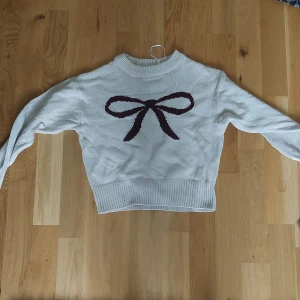 Vit stickad tröja med rosett - Superfin vit stickad tröja med en stor mörklila rosett framtill. Tröjan har rund halsringning, långa ärmar och ribbade muddar vid ärmslut och nederkant. Perfekt för en söt och trendig look. Helt ny!