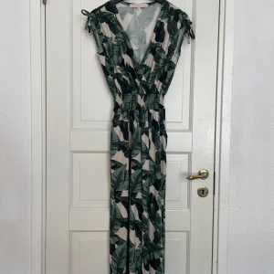 Grönmönstrad jumpsuit med knytband - Säljer en jumpsuit med tropiskt bladmönster i grönt och beige. Den har v-ringning, resår i midjan och knytband på axlarna. Perfekt för sommardagar eller semester. Luftigt material och snygg passform.