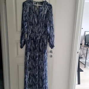Blåvit långklänning med mönster från H&M - Säljer en blåvit långklänning från H&M med abstrakt mönster och lång ärm. Klänningen har en snygg detalj med guldfärgad ring vid halsen och en lätt, luftig känsla. 