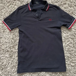 Svart pikétröja från Fred Perry - Säljer en svart pikétröja från Fred Perry med klassisk krage och knäppning framtill. Tröjan har röda och vita ränder på krage och ärmslut samt röd broderad logga på bröstet. Perfekt för en stilren och sportig look.