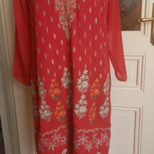 Röd klänning med broderade detaljer från Rupali - Säljer en röd klänning från Rupali med vackra broderade mönster i vitt och orange längs nederkanten och framtill. Klänningen har långärmade transparenta ärmar och raka linjer. Perfekt för dig som vill sticka ut med färg och mönster.