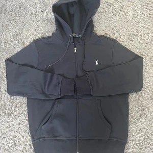 Mörk blå hoodie med zip Ralph Lauren - STORLEK S. Snygg mörkblå hoodie från Ralph Lauren med dragkedja och luva. Tröjan har två fickor framtill och broderade loggan på bröstet. Perfekt för en avslappnad och lyxig stil. Helt oanvänd och helt ren. Jätte bra material och bekväm. PRISET KAN DISKUTERAS.