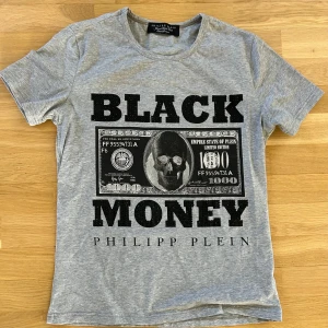 Grå t-shirt med tryck från Philipp Plein - Snygg grå t-shirt från Philipp Plein med stort svart tryck 'BLACK MONEY' och dollar-motiv framtill. T-shirten har korta ärmar och är dekorerad med små svarta nitar på texten. Rakt snitt och klassisk rund hals. Väldigt liten i storlek. Står L men är som S