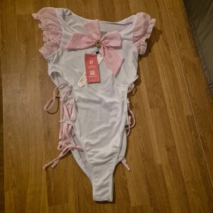 Vit och rosa cosplay-body med rosett från Romwe - Supersöt body i vitt med rosa volangärmar och stor rosa rosett framtill. Snygga knytband på sidorna och dekorativ detalj i mitten av rosetten. Perfekt för cosplay! Storlek M  Oanvänd med orginallappen kvar. Se även mina andra auktioner med möjlighet till bundles med rabatt.