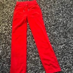 Röda velourbyxor från Juicy Couture - Säljer ett par klassiska röda velourbyxor från Juicy Couture med snörning i midjan och fickor både fram och bak. Byxorna har broderad logga på ena fickan och är perfekta för en avslappnad stil.