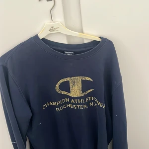 Blå sweatshirt från Champion - Mörkblå sweatshirt från Champion med stort gult logotryck och texten 'Champion Athleticwear Rochester, N.Y.' på bröstet. Tröjan har rund halsringning och långa ärmar. Perfekt för en avslappnad stil.