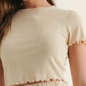 Beige croppad topp från NA-KD - Säljer en beige croppad topp från NA-KD med korta ärmar och vågig kant vid ärmslut och nederkant. Toppen är ribbad och har en rund halsringning. Perfekt för en enkel och stilren look.
