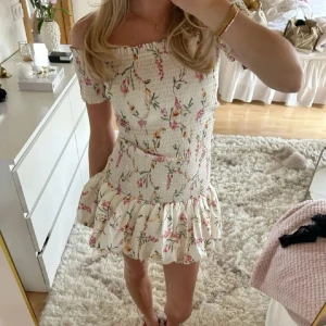 Blommig offshoulder klänning med volang - Supersöt kortklänning med blommigt mönster och offshoulder-modell. Klänningen har volang nertill och är perfekt för sommarens alla tillfällen. 
