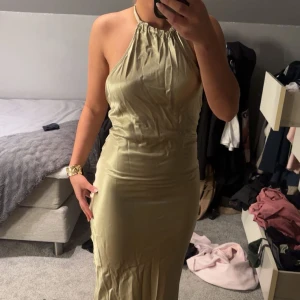 Guldig långklänning med halterneck - Säljer en elegant guldig balklänning med halterneck och öppen rygg. Klänningen har en slät, glansig yta och knyts i nacken. Perfekt för speciella tillfällen när du vill sticka ut.