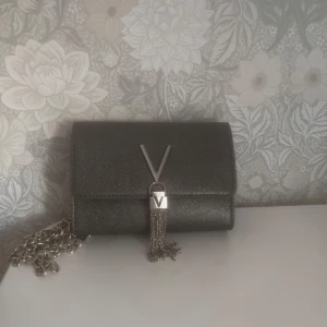 Svart glittrig axelväska med kedja, valentino - Säljer en svart glittrig axelväska från Valentino med silverfärgad kedja och V-detalj framtill. Väskan har en elegant design och är perfekt för att lyfta din outfit. Liten och smidig storlek, passar till det mesta.