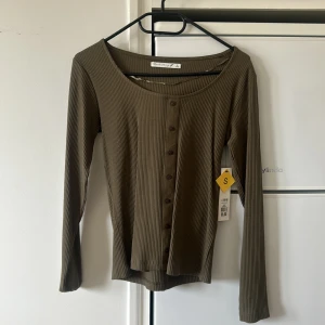 Olivgrön ribbad topp med knappar  - Säljer en olivgrön ribbad topp med långa ärmar och knappar framtill. Toppen har en rund halsringning och är tillverkad i ett stretchigt material som sitter snyggt på kroppen. Perfekt att styla till jeans eller kjol.