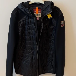 Svart hybridjacka från Parajumpers - Snygg svart jacka från Parajumpers med quiltad front och ärmar i stickad struktur. Jackan har huva, dragkedja och en gul detalj vid halsen. Märkespatch på ena ärmen. Perfekt för kyliga dagar.