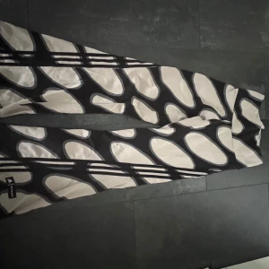 Svarta och vita mönstrade byxor från Adidas x Marimekko - Unika byxor från Adidas i samarbete med Marimekko, med ett stort svartvitt grafiskt mönster. Byxorna har normal passform och är tillverkade i ett mjukt syntetmaterial. Perfekta för dig som vill sticka ut med en cool design.