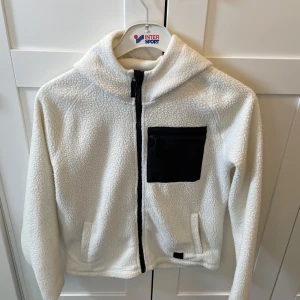 Vit fleecejacka med huva från McKinley - Mysig vit fleecejacka från McKinley med svart dragkedja och svart bröstficka. Jackan har huva och två sidofickor, perfekt för kyliga dagar. Enkel och stilren design som passar till mycket.