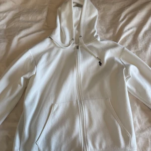 Vit zip hoodie från Polo Ralph Lauren - Säljer en klassisk vit hoodie från Polo Ralph Lauren med dragkedja och huva. Jackan har två fickor framtill och en broderad logga på bröstet. Perfekt för en stilren och avslappnad look. Knappt använd då den inte riktigt matchade min stil