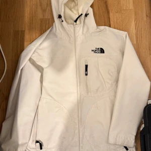 Vit vindjacka från The North Face - Säljer en vit vindjacka från The North Face med huva och dragkedja framtill. Jackan har en bröstficka med dragkedja och logga på både fram- och baksidan. Perfekt för blåsiga dagar och enkel att matcha. Jätte bra skick.
