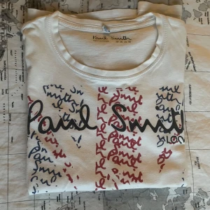 Vit t-shirt med Union Jack och bil från Paul Smith - Vit t-shirt från Paul Smith med Union Jack-flagga i rött och blått, handskrivna loggor och en ikonisk randig bil på framsidan. T-shirten är i 100% bomull och har rund hals och korta ärmar. 