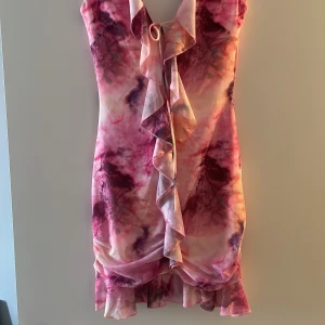 Rosa och lila volangklänning med knyt - Säljer en färgstark kortklänning i rosa och lila toner med volangdetaljer framtill och knyt i halsen. Klänningen har ett unikt mönster och öppen rygg, perfekt för dig som vill sticka ut. Materialet känns mjukt och följsamt.