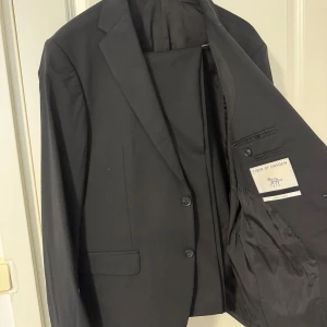 Svart kostym från Tiger of Sweden - Klassisk svart kostym från Tiger of Sweden med kavaj och matchande byxor. Kavajen har två knappar, smala slag och innerficka. Byxorna är raka och stilrena. Perfekt för studenten.