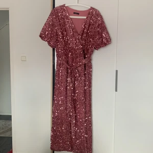 Rosa paljettklänning från SHEIN - Glittrig rosa klänning från SHEIN med omlottmodell och knytband i midjan. Klänningen har korta ärmar och är täckt av paljetter som ger en festlig look. Perfekt för dig som vill sticka ut på festen!
