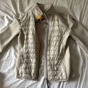 Beige jacka från Parajumpers warm up  - Snygg beige jacka från Parajumpers med quiltade partier framtill och på axlarna. Jackan har lång ärm, hög krage med gul detalj och dragkedja framtill. Perfekt för övergångssäsonger och har en sportig, stilren look.