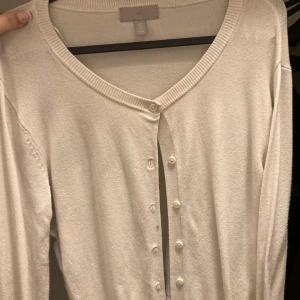Vit kofta från H&M - Säljer en klassisk vit kofta från H&M i storlek M. Koftan har långa ärmar, rund halsringning och knäppning framtill med matchande vita knappar. Perfekt att slänga över en topp eller klänning för en enkel och stilren look.