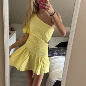 Gul one shoulder klänning med volang - Säljer en söt gul kortklänning med one shoulder-design och volang nertill. Klänningen har en asymmetrisk axel och en liten cut-out i midjan. Den är helt ny och helt oanvänd!