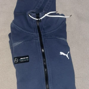 Blå hoodiejacka från Puma x Mercedes AMG - Mörkblå hoodiejacka med dragkedja från Puma i samarbete med Mercedes AMG Petronas. Jackan har vit Puma-logga på bröstet, svart badge med Mercedes AMG och vit dragsko i huvan. Perfekt för dig som gillar motorsport och streetwear.