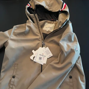 Beige vindjacka från Moncler med huva - Säljer en beige vindjacka från Moncler med huva och dragkedja. Jackan har svarta detaljer, två fickor framtill och Moncler-logga på ärmen. Insidan är svart med Moncler-mönster och serieruta. Röd och vit rand på huvan. Storlek M men passar även S