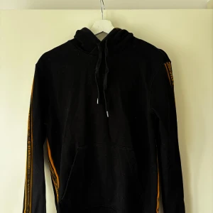 Svart hoodie från G-Star RAW med gula detaljer - Svart hoodie från G-Star RAW med huva och dragsko. Gula ränder med text längs ärmarna ger en sportig look. Klassisk känguruficka framtill och ribbade muddar. Perfekt för en avslappnad stil.